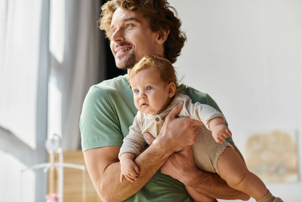 кудрявый и довольный отец держит в руках своего маленького сына в уютной спальне дома, отцовство - Фото, изображение