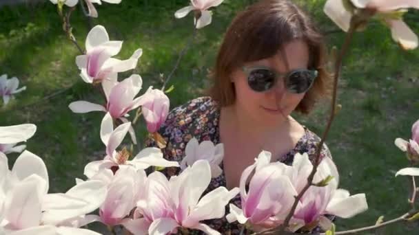 Happy woman in sunglasses enjoying nature and touching blooming magnolia flowers in the garden - 映像、動画