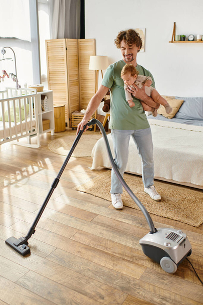 happy father multitasking housework and childcare, man vacuuming bedroom with infant son in arms - Photo, Image