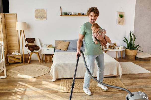 gelukkig man multitasking huishoudelijk werk en kinderopvang, vader stofzuigen slaapkamer met zuigeling zoon in armen - Foto, afbeelding