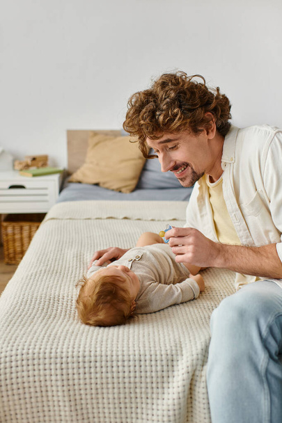 kudrnatý a vousatý otec držící dudlík poblíž kojeneckého chlapečka na posteli, otcovství a lásce - Fotografie, Obrázek