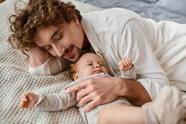 szczęśliwy mężczyzna z kręconymi włosami obejmujący swoje niemowlę leżąc razem na łóżku, pielęgnując i kochając - Zdjęcie, obraz