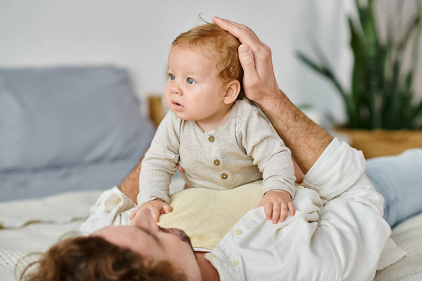 uomo con i capelli ricci che abbraccia il figlio neonato con gli occhi azzurri su un letto, legando tra padre e figlio - Foto, immagini