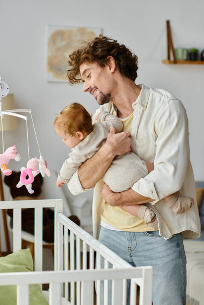 glimlachende vader met krullend haar en baard houden zijn baby zoon in de buurt van witte baby wieg thuis - Foto, afbeelding