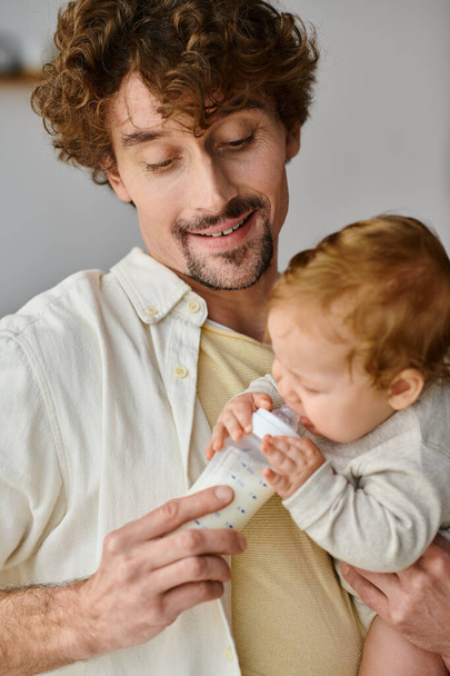赤ん坊のボトルから栄養価の高いミルクを授乳するひげを持つ注意深い父親,養育 - 写真・画像