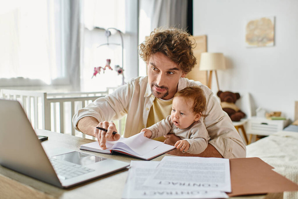 man holding in arms his infant son while working remotely on laptop from home, work-life balance - Photo, Image