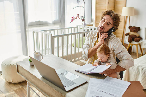 man holding in arms infant son while talking on smartphone and working from home, work-life balance - Photo, Image