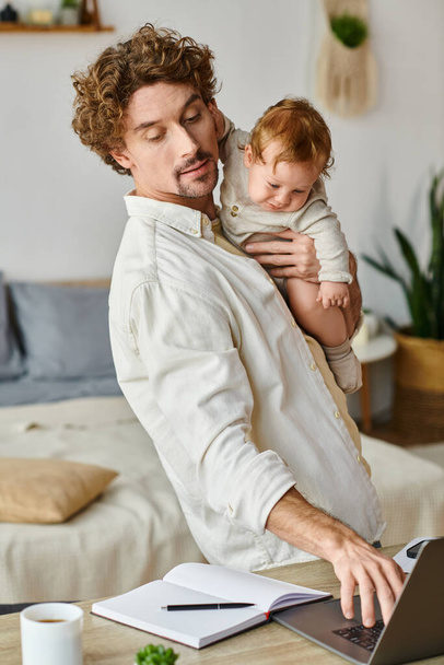 pai feliz e encaracolado segurando nos braços seu filho bebê e usando laptop na mesa, equilíbrio de vida no trabalho - Foto, Imagem