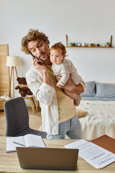 egyedülálló apa gazdaság csecsemő fia, miközben beszél okostelefon közelében laptop az asztalon, távoli munka - Fotó, kép