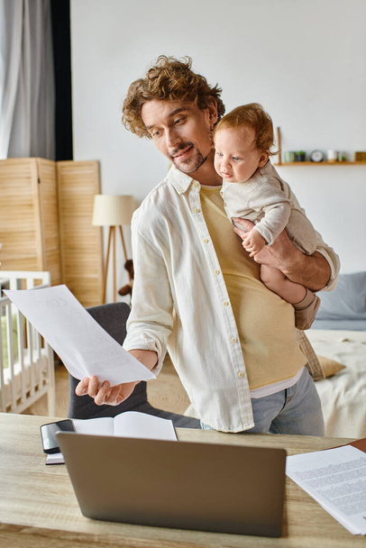 happy and bearded father holding in arms infant child and looking at document, work life balance - Photo, Image