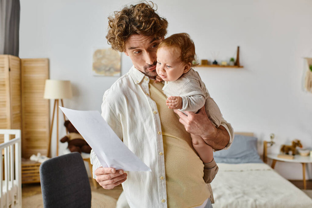 lockiger und bärtiger Vater hält Säugling im Arm und betrachtet Dokument, Work-Life-Balance - Foto, Bild