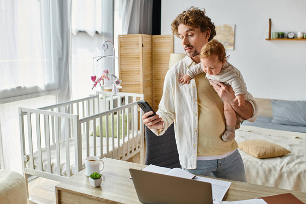 homem barbudo com cabelo encaracolado segurando filho bebê e usando smartphone perto de laptop, trabalhar em casa - Foto, Imagem