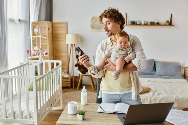 elfoglalt egyedülálló apa gazdaság csecsemő fia és használja okostelefon közelében laptop és cumisüveg az asztalon - Fotó, kép