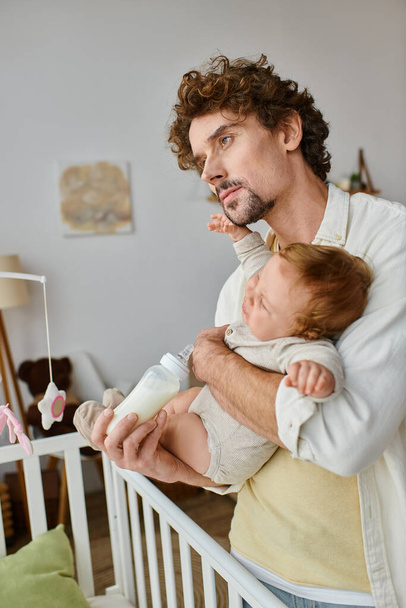 Kıvırcık saçlı baba, bebeğini beşik, babalık ve bakım yakınlarındaki bir şişeden besliyor. - Fotoğraf, Görsel