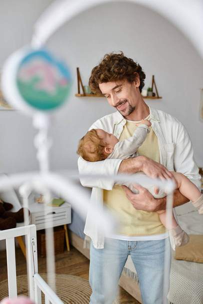 pai solteiro feliz segurando nos braços seu filho bebê perto berço no berçário, paternidade e amor - Foto, Imagem