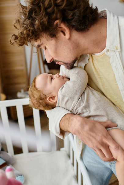 заботливый отец-одиночка, поднимающий своего маленького сына из детской кроватки в детской, отцовство и любовь - Фото, изображение