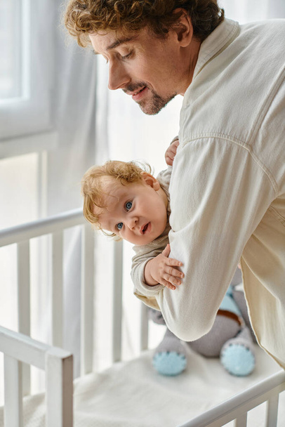 pai amoroso e feliz levantando seu filho bebê com olhos azuis do berço do bebê no berçário, paternidade - Foto, Imagem