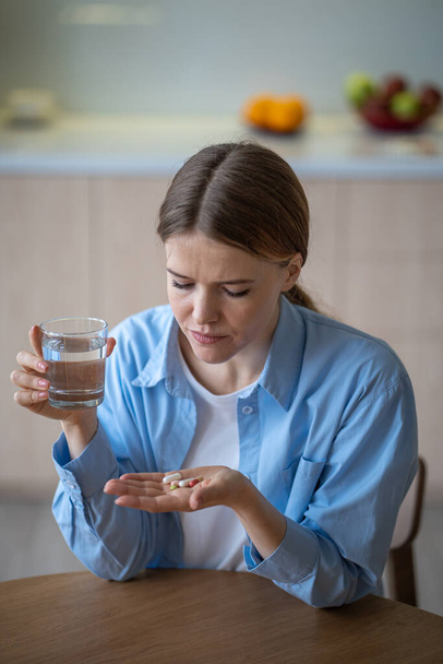 Mujer que sufre depresión tomando antidepresivos neurolépticos beber agua sentada en la cocina en casa. Mujer triste tratando con pastillas, tabletas. Enfermedad mental, trastorno de personalidad, enfermedad endógena - Foto, imagen