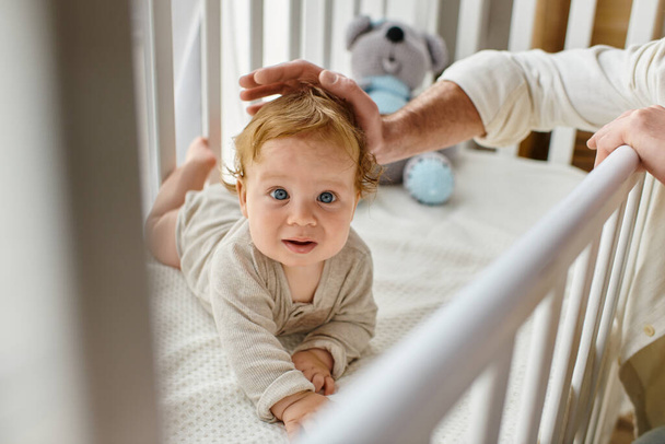 vue recadrée de l'homme caressant les cheveux de bébé garçon aux yeux bleus dans la crèche, main du père près de l'enfant - Photo, image