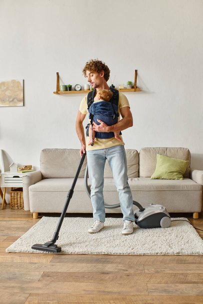 vader met zuigeling baby jongen in drager stofzuigen appartement thuis, netheid en huishoudelijk werk - Foto, afbeelding