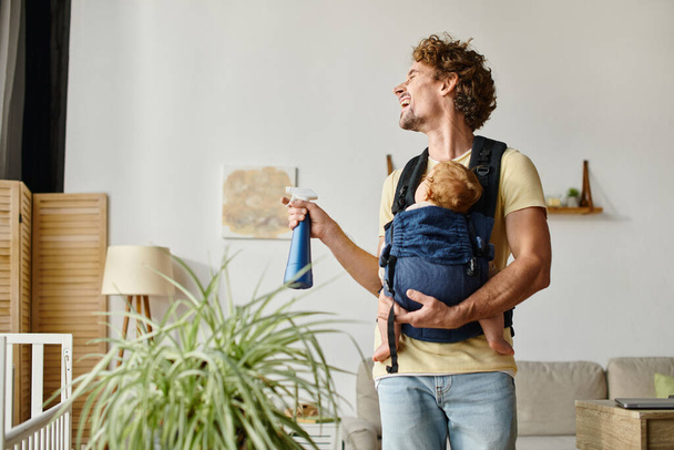 šťastný otec s kojencem syn v nosiči drží sprej láhev při smíchu a zalévání zelená rostlina - Fotografie, Obrázek