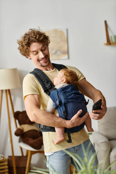 curly haired and happy man messaging on smartphone while baby asleep in carrier, modern parenting - Photo, Image