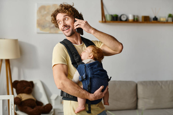 cheerful man talking on smartphone with baby sleeping in carrier, balance between life and work - Photo, Image