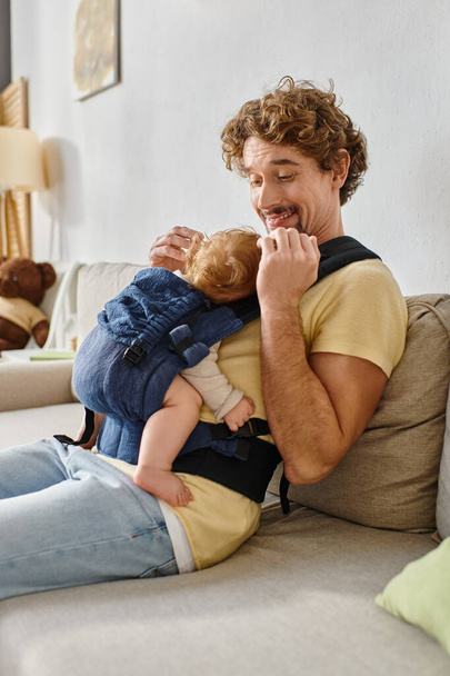 allegro padre dai capelli ricci seduto sul divano con figlio neonato in marsupio, paternità e amore - Foto, immagini