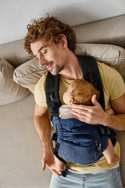 górny widok zadowolony ojciec siedzi na kanapie z niemowlęcym synem w nosidełku, ojcostwo i miłość - Zdjęcie, obraz