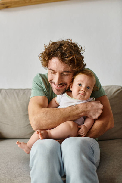 fröhlicher Vater mit Bart umarmt seinen kleinen Sohn mit blauen Augen in einem gemütlichen Wohnzimmer, Porträt - Foto, Bild