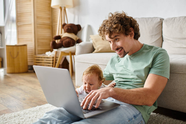 wesoły ojciec za pomocą laptopa w pobliżu niemowlęcia syna w salonie, równoważąc między ojcostwem a pracą - Zdjęcie, obraz