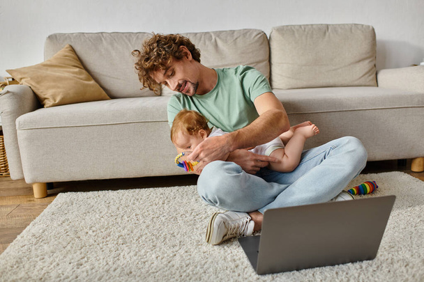 glücklicher Mann mit lockigem Haar hält Babyrassel in der Nähe von Säugling und Laptop, Work-Life-Balance - Foto, Bild
