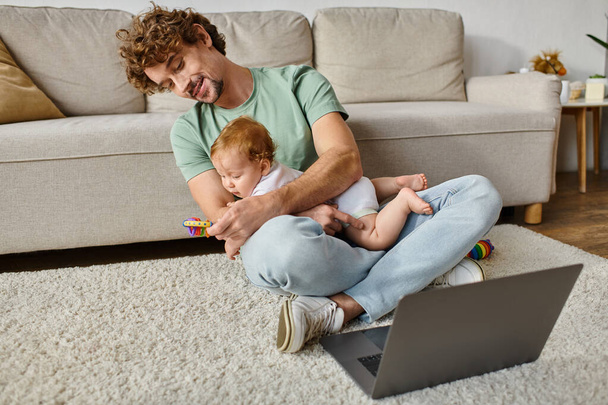 Χαρούμενος άντρας με σγουρά μαλλιά κρατώντας κουδουνίστρα μωρού κοντά σε νεογέννητο γιο και φορητό υπολογιστή, εργασία και ισορροπία ζωής - Φωτογραφία, εικόνα