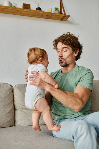 padre sosteniendo en brazos lindo bebé hijo y tener un momento feliz en el sofá en la sala de estar - Foto, Imagen