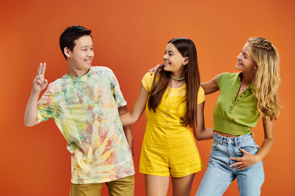 fröhliche multikulturelle Teenager in lebhaften lässigen Outfits, die sich auf orangefarbenem Hintergrund anschauen - Foto, Bild