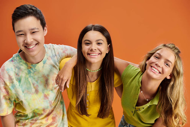 vreugdevolle multiraciale tieners in casual kleding kijken naar de camera op oranje achtergrond, vriendschap dag - Foto, afbeelding