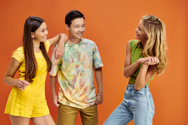 αξιολάτρευτο ευχάριστα ποικίλες έφηβοι σε ζωντανή ενδυμασίες κοιτάζοντας ο ένας τον άλλον σε πορτοκαλί φόντο - Φωτογραφία, εικόνα