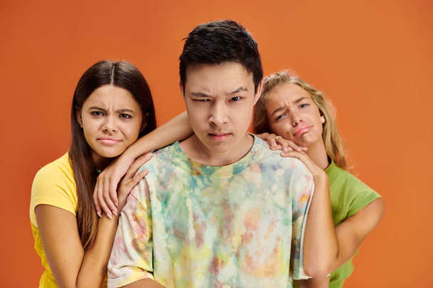 Verwirrte Teenager in grellen Kostümen, die vor orangefarbener Kulisse in die Kamera schauen, Freundschaftstag - Foto, Bild