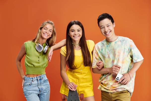три веселых подростка с наушниками, скейтбордом и смартфоном смотрят в камеру, дружба - Фото, изображение