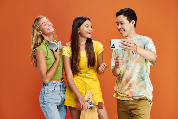 radostné rozmanité dospívající s sluchátky a skateboard přičemž selfie na oranžovém pozadí, přátelství den - Fotografie, Obrázek
