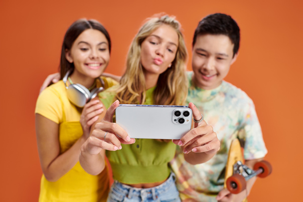 focus on smartphone in hands of three jolly blurred diverse friends taking selfie on orange backdrop - Photo, Image