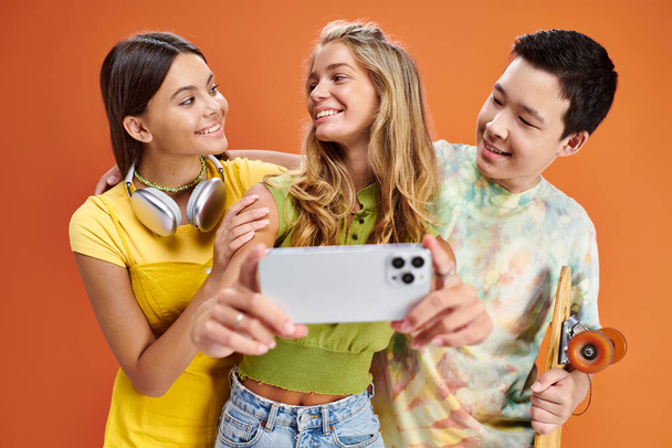 radostné rozmanité přátele v živých oděvech přičemž selfie a usmívat se na sebe na oranžovém pozadí - Fotografie, Obrázek