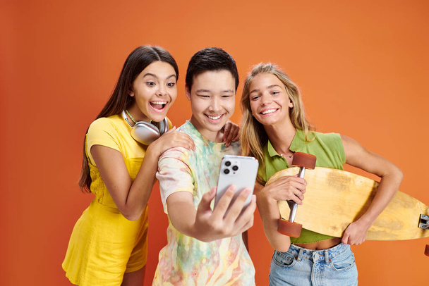 joyeux amis adolescents multiraciaux en vêtements vifs avec écouteurs et planche à roulettes prendre selfie - Photo, image
