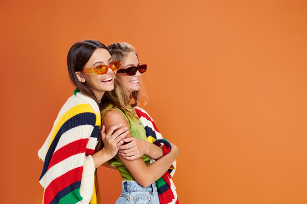 jolly pretty teenage friends with sunglasses hugging and covering with blanket on orange backdrop - Photo, Image