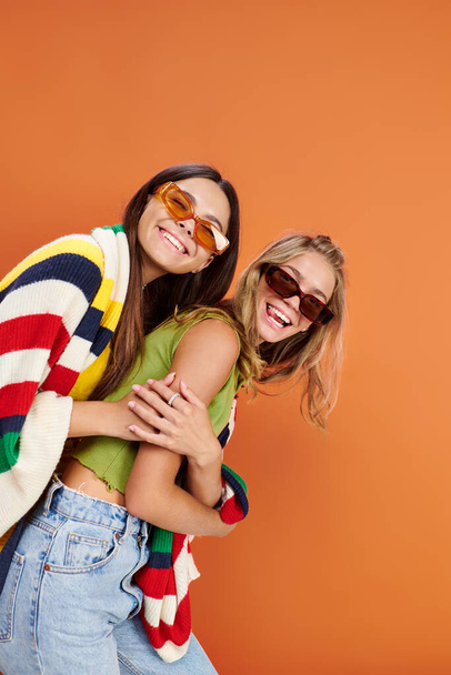 fröhliche entzückende Teenager-Freunde mit Sonnenbrille, die sich umarmen und gemeinsam vor orangefarbener Kulisse Spaß haben - Foto, Bild