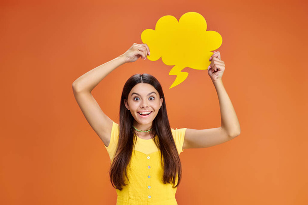 cheerful adolescent girl in everyday outfit with thought bubble looking at camera on orange backdrop - Photo, Image