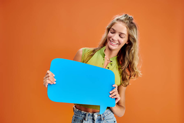 vidám szőke kamasz lány pózol kék gondolat buborék a kezében narancssárga háttér - Fotó, kép
