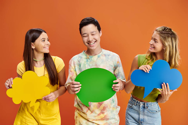 vreugdevolle adolescent interraciale vrienden houden spraak en gedachte bubbels op oranje achtergrond - Foto, afbeelding