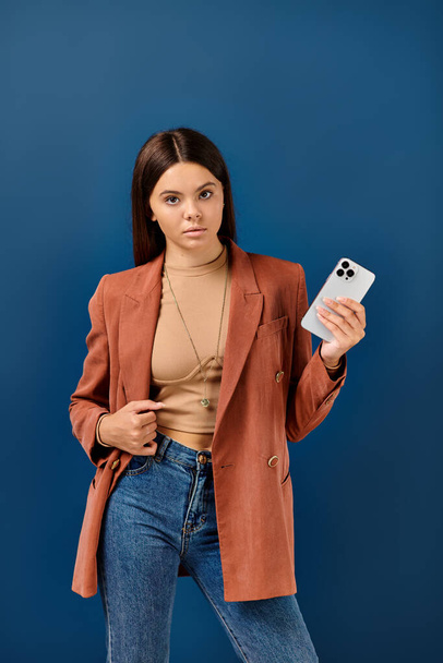 стильная девушка-подросток в элегантном коричневом блейзере, позирующая с мобильным телефоном в руке и смотрящая в камеру - Фото, изображение