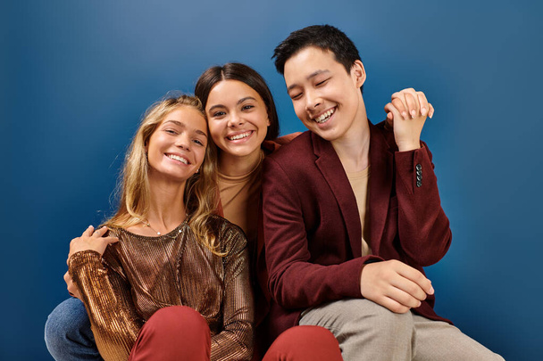 alegre adolescente niñas sonriendo en cámara al lado de su alegre elegante asiático amigo en azul fondo - Foto, Imagen
